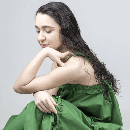 Modella con abito verde, capelli lunghi e mossi, in posa contemplativa con il braccio appoggiato sul ginocchio.