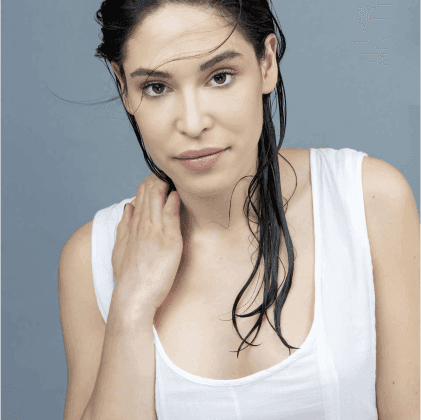 Ritratto di una donna con capelli bagnati, indossando una maglietta bianca, con uno sfondo grigio chiaro.
