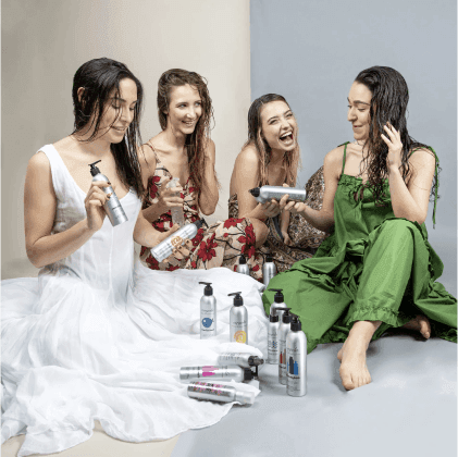 Quattro donne con capelli bagnati che tengono prodotti per capelli in bottiglie grigie, indossando abiti colorati e sorridendo.