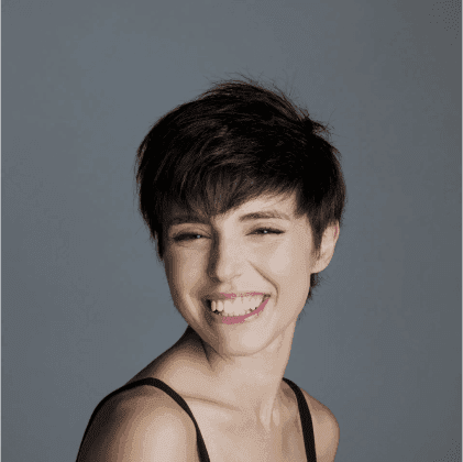 Ritratto di una giovane donna con capelli corti e scuri, sorridente, su uno sfondo grigio chiaro.