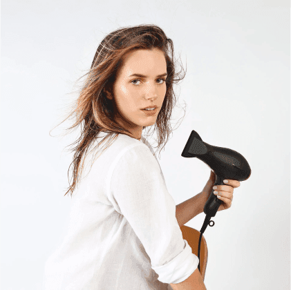 Modella con capelli mossi che tiene un asciugacapelli nero, indossando una camicia bianca, in un ambiente neutro.