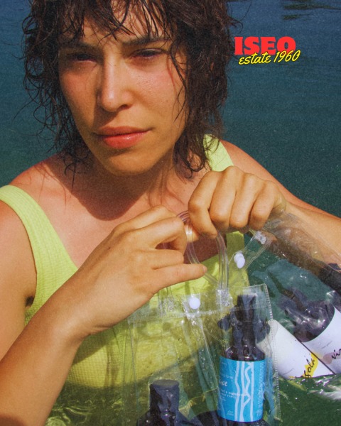 Donna con capelli ricci e bagnati che tiene in mano bottiglie di prodotti per capelli, indossando un top giallo chiaro.