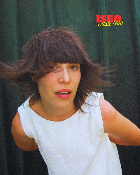 Donna con capelli mossi e abito bianco, posa con espressione naturale su sfondo verde scuro, logo ISEO in alto a destra.