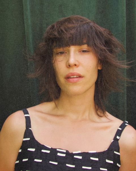 Donna con capelli disordinati e taglio corto, indossa un abito a righe bianche e nere. Espressione neutra e sguardo diretto verso la fotocamera.