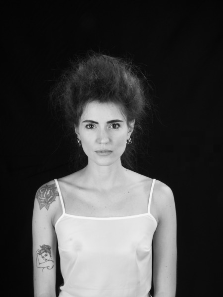Ritratto di una donna con capelli voluminosi e un tatuaggio sul braccio, indossando una canotta bianca su sfondo nero.