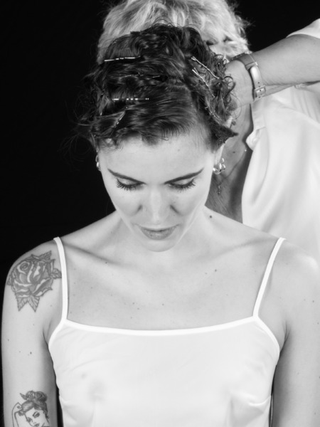 Donna con capelli ricci e fermagli, mentre un'altra persona le sistema i capelli. Mostra un look elegante e curato.