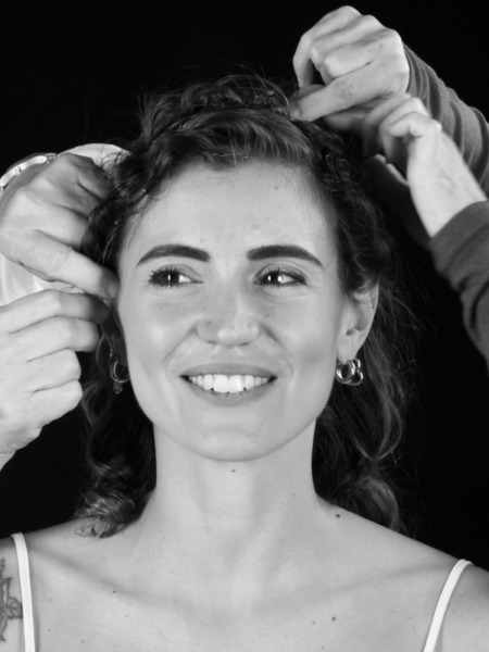 Donna con capelli ricci mentre viene acconciata da due persone, sorridendo verso la fotocamera.