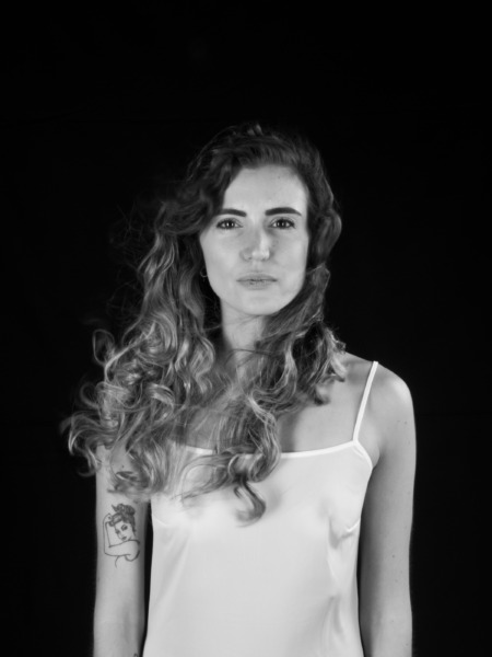 Donna con capelli ricci e lunghi, indossando una canotta bianca, in un ritratto su sfondo nero.